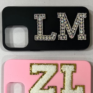 Puede incluir: Dos fundas para teléfono con diseños diferentes. La primera funda es negra con las letras "LM" en pedrería. La segunda funda es rosa con las letras "ZL" en blanco con ribetes dorados.