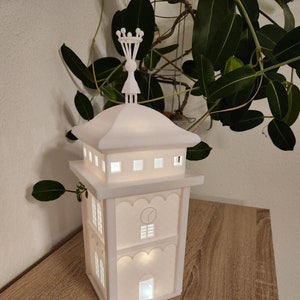 Könnte beinhalten: Eine weiße, beleuchtete, turmförmige dekorative Lampe. Die Lampe hat ein mehrstufiges Design mit Fenstern und einer dekorativen Oberseite. Die Lampe steht auf einer Holzoberfläche, mit grünem Laub im Hintergrund.