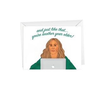 May include: A greeting card with a cartoon illustration of a woman in a green robe sitting in front of a laptop. The text on the card reads "and just like that... you're another year older!"
