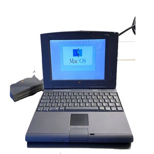 Könnte beinhalten: Ein schwarzer Apple PowerBook 1400 Laptop mit einem integrierten Trackpad und einem schwarzen externen Diskettenlaufwerk. Der Laptop-Bildschirm zeigt den Systemstartbildschirm von Mac OS an.