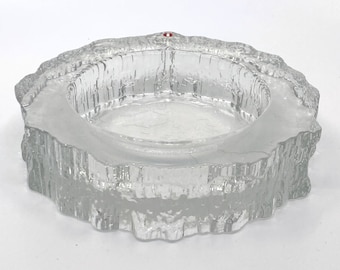 May include: A clear glass candle holder with a textured, icicle-like design. The holder has a round shape and a small red dot on the top.