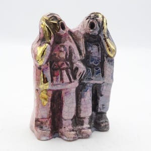May include: Two ceramic figurines of people with their hands over their ears, one in pink and gold and the other in blue and white.