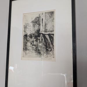 May include: Black framed print of a detailed street scene. The artwork, in monochrome, depicts buildings and a person in a narrow alleyway. The print is mounted with a white mount and framed in a black wooden frame.