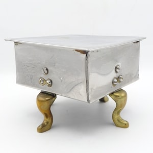 May include: A silver metal box with a rectangular form and a slightly overhanging lid. The box is supported by four brass-coloured, curved legs. The box has visible rivets and a simple, classic design.