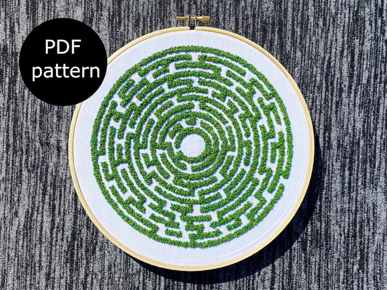 May include: A white embroidery hoop with a green embroidered maze pattern. The maze is a circular design with a smaller circle in the center.