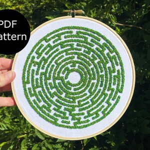 May include: A white embroidery hoop with a green and blue embroidered maze pattern. The pattern is a circular maze with a central circle.