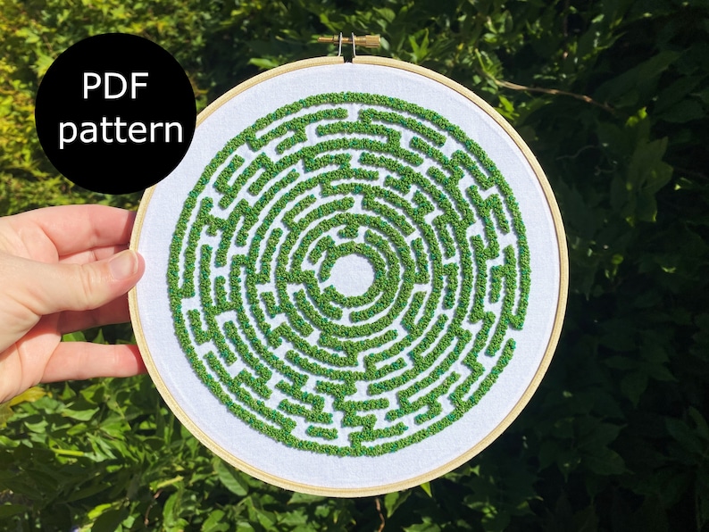 May include: A white embroidery hoop with a green and blue embroidered maze pattern. The pattern is a circular maze with a central circle.