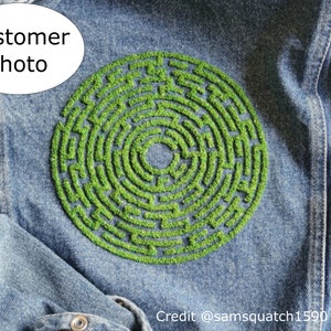 May include: A denim jacket with a green embroidered circular maze design. The maze is made of small green stitches that create a raised effect.