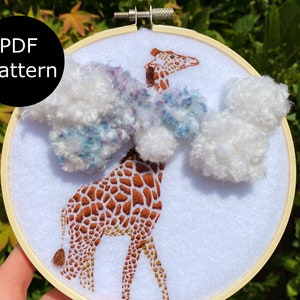 Puede incluir: Un aro de bordado con una jirafa bordada en marrón y blanco con pompones blancos y azules. El aro es de madera y tiene un fondo de tela blanco. La jirafa está bordada con hilo marrón y blanco. Los pompones son de hilo y están unidos al aro con una aguja e hilo. El aro tiene un diámetro de 15 cm.