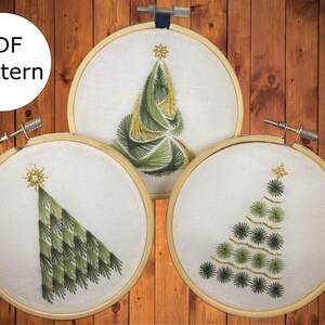 May include: Three embroidery hoops featuring different Christmas tree designs. The trees are embroidered in green and gold thread on white fabric.