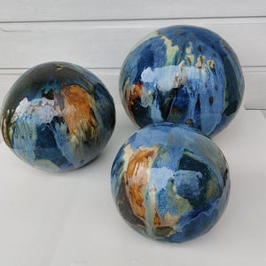 Könnte beinhalten: Drei glasierte Keramikkugeln in verschiedenen Größen. Die Kugeln zeigen eine Mischung aus blauen, braunen und cremefarbenen Farbtönen mit marmoriertem Aussehen. Das Arrangement besteht aus einer großen, einer mittleren und einer kleinen Kugel, jede mit einzigartigen Mustern.