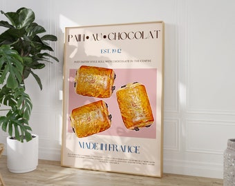 May include: A framed art print featuring the words "PAIN AU CHOCOLAT" and "MADE IN FRANCE". The print displays three golden-brown puff pastry rolls with chocolate filling against a pink background. The print is in a light-colored wooden frame.