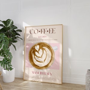 May include: A poster with a pink background and a gold frame. The poster features a stylized illustration of a cup of coffee with latte art. The text on the poster reads "COFFEE EST. 1979 BREWED FRESHLY IN STORE FROM ROASTED COFFEE BEANS MADE IN ITALY".