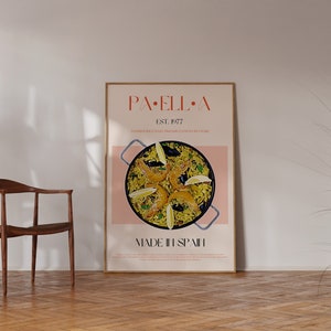 May include: A framed poster with a pink background and a photo of a paella dish with shrimp, mussels, and lemon wedges. The poster has the text "PAELLA" in a red font and "EST. 1977" in a smaller font. The text "MADE IN SPAIN" is at the bottom of the poster.