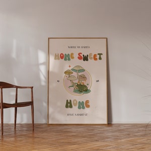 May include: A framed poster with the words "HOME SWEET HOME" in colorful, retro-style lettering. The poster features an illustration of mushrooms and a frog. The text "WHERE MY HABITS HAVE A HABITAT" is also included.