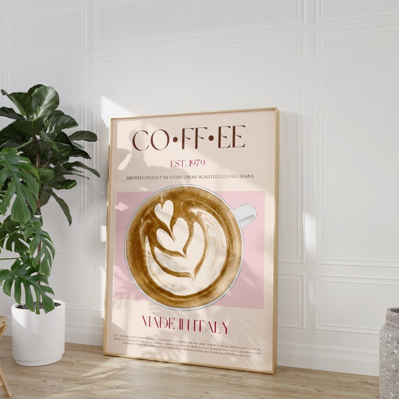 May include: A poster with a pink background and a gold frame. The poster features a stylized illustration of a cup of coffee with latte art. The text on the poster reads "COFFEE EST. 1979 BREWED FRESHLY IN STORE FROM ROASTED COFFEE BEANS MADE IN ITALY".