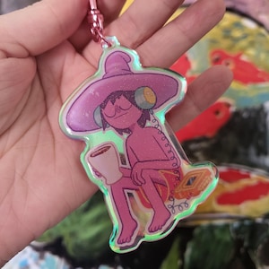 May include: A pink iridescent keychain featuring a cartoon character wearing a purple hat and holding a cup of coffee. The character has pink skin and is wearing headphones. The keychain is attached to a silver chain.