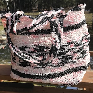 May include: A large, striped tote bag made from crocheted plastic. The bag is black, white, and pink with a striped pattern. The bag has two long handles.