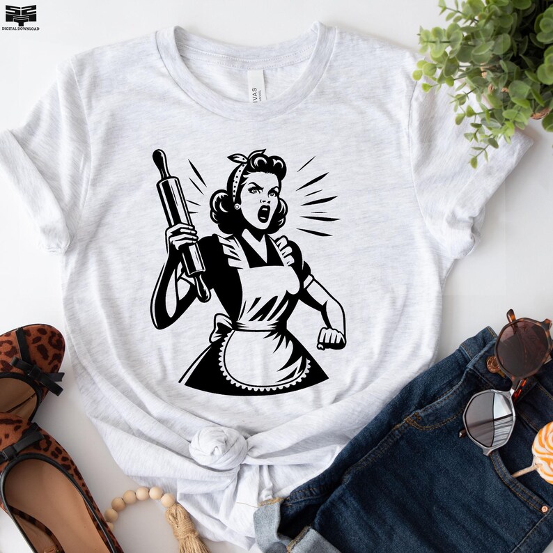 May include: A white t-shirt with a black and white illustration of a woman in a dress and apron holding a rolling pin. The woman is yelling and has a determined expression on her face.