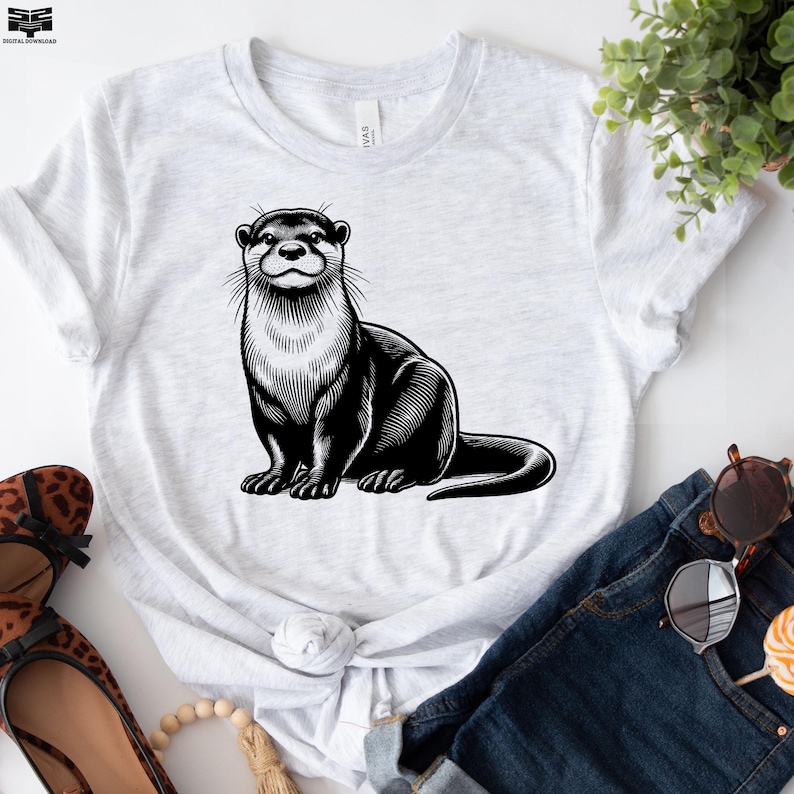Puede incluir: Una camiseta blanca con una ilustraci&oacute;n en blanco y negro de una nutria. La nutria est&aacute; sentada sobre sus patas traseras con sus patas delanteras descansando sobre su pecho. La nutria tiene un cuerpo largo y delgado y una cola tupida.