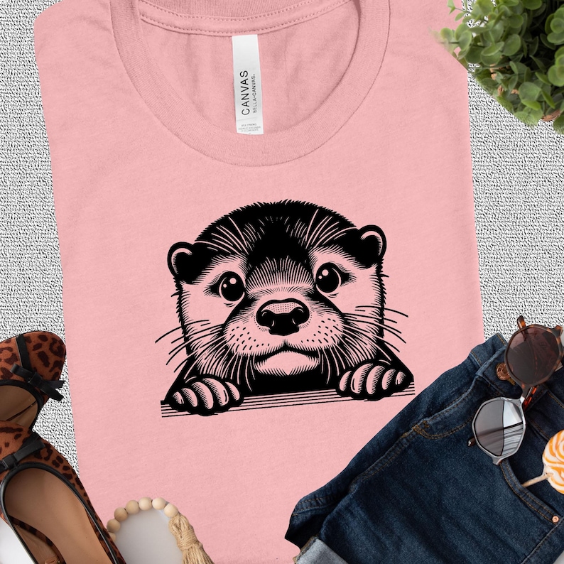 Puede incluir: Una camiseta rosa con una ilustraci&oacute;n en blanco y negro de una nutria asom&aacute;ndose por el borde de la camiseta. La nutria tiene una expresi&oacute;n linda y de tipo caricatura.