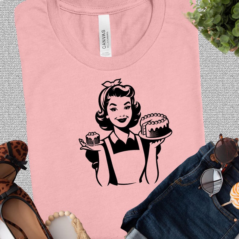May include: A pink t-shirt with a black and white illustration of a woman holding a cake and a cupcake. The woman is wearing a dress and an apron.