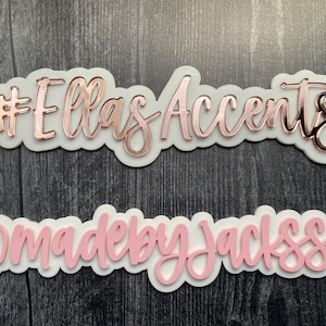 May include: Two decorative signs with text. The top sign reads "#EllasAccents" in rose gold script over a white base. The bottom sign reads "@madebyjacksss" in pink script over a white base.