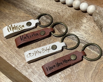 May include: Four personalized leather keychains in white and brown with gold-colored key rings. Each keychain has a name or message engraved on it: "Melissa", "Karina", "Mrs. Balagh", and "I love you Mom".