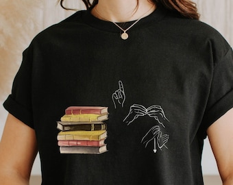 May include: A black t-shirt with a white graphic of a stack of books and two hands signing the word "book" in American Sign Language.