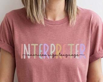 May include: A woman wearing a maroon t-shirt with the text "INTERPRETER" in rainbow colors and the text "american sign language" in black below it.