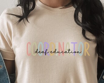 Może przedstawiać: Beżowa koszulka z napisem "Coordinator deaf education" w kolorach tęczy.