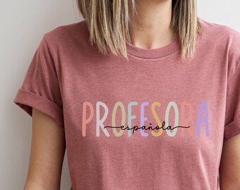 Może przedstawiać: Kobieta w bordowym t-shircie z napisem "PROFESORA -española-  " w kolorowych literach. Ma na sobie niebieskie dżinsy i siedzi ze skrzyżowanymi nogami.