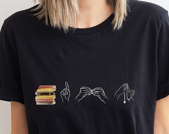 Może przedstawiać: Czarny T-shirt z białym nadrukiem stosu książek i słowem "CZYTAJ" w języku migowym.