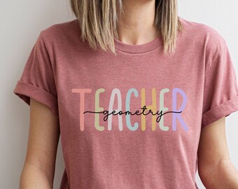Może przedstawiać: Różowy T-shirt z napisem "TEACHER geometry" w kolorach tęczy.