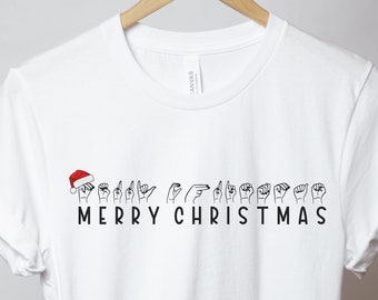 May include: White t-shirt with the words "Merry Christmas" in black text and a row of hands signing the phrase in American Sign Language. A red Santa hat is on top of the first hand.
