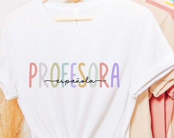 Może przedstawiać: Biała koszulka z napisem "PROFESORA española" w kolorach tęczy. Koszulka wisi na drewnianym wieszaku.
