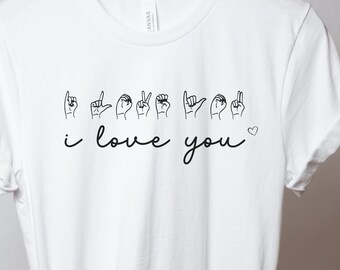 May include: White t-shirt with the phrase "i love you" written in black lettering. The phrase is signed in American Sign Language.