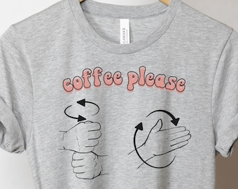 May include: Gray t-shirt with the text "coffee please" in pink letters. The text is in a retro font. Two hand gestures are shown below the text. The first gesture is a hand making a circular motion with the fingers pointing up. The second gesture is a hand making a circular motion with the palm facing out.