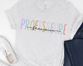 Może przedstawiać: Jasnoszary t-shirt z napisem "PROFESSEURE française" w kolorach tęczy. Koszulka jest złożona i leży płasko na białym tle.