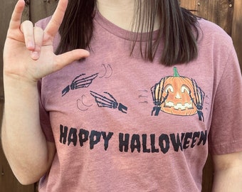 May include: A mauve t-shirt with a graphic of a pumpkin with hands covering its eyes and the text "Happy Halloween".