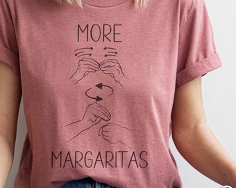 May include: A pink t-shirt with the text "MORE MARGARITAS" in black. The text is accompanied by a graphic of hands signing the phrase in American Sign Language.