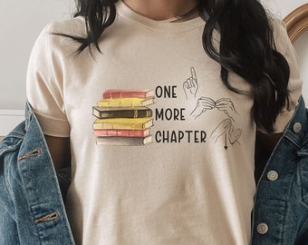 May include: A beige t-shirt with a stack of books and the text "ONE MORE CHAPTER" in black. The text is accompanied by a hand sign for the letter "C" in American Sign Language.