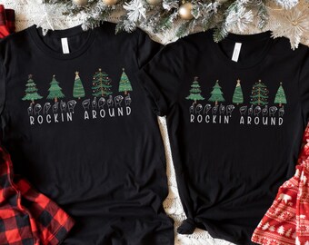 May include: Two black t-shirts with a graphic of Christmas trees and the words "Rockin' Around" in sign language. The shirts are laid out on a white fuzzy surface with red and white plaid fabric and a Christmas tree.