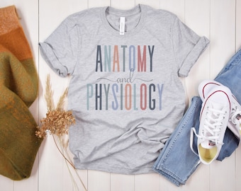 Może przedstawiać: Szary T-shirt z napisem "Anatomia i fizjologia" w kolorowych literach. Tekst jest w tęczowej kolorystyce.