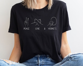 Może przedstawiać: Czarny T-shirt z białym nadrukiem graficznym gestów dłoni i tekstem "Peace Love & Hornets".