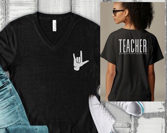 Może przedstawiać: Czarny T-shirt z dekoltem w kształcie litery V z białym gestem „Kocham cię” w języku migowym na lewej piersi. Z tyłu koszulki napis „TEACHER of deaf and hard of hearing” białym kolorem.