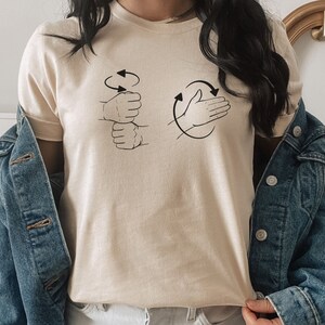 May include: A cream colored t-shirt with a graphic of two hands. One hand is making a fist and the other hand is open with the palm facing out. Both hands have arrows circling them.