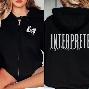 以下が含まれることがあります： 黒色のジッパー付きフード付きスウェットシャツ。背面に「INTERPRETER sign language」の文字、前面に手話記号がプリントされています。