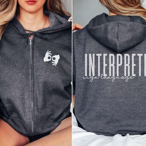以下が含まれることがあります： ジッパー付きのダークグレーのフード付きスウェットシャツ。背面には「INTERPRETER sign language」の文字、前面には手話記号が描かれています。