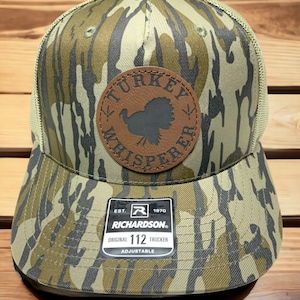 May include: A camouflage trucker hat with a brown leather patch that says "Turkey Whisperer" and a silhouette of a turkey. The hat is adjustable and has the Richardson logo and "112" printed on the back.
